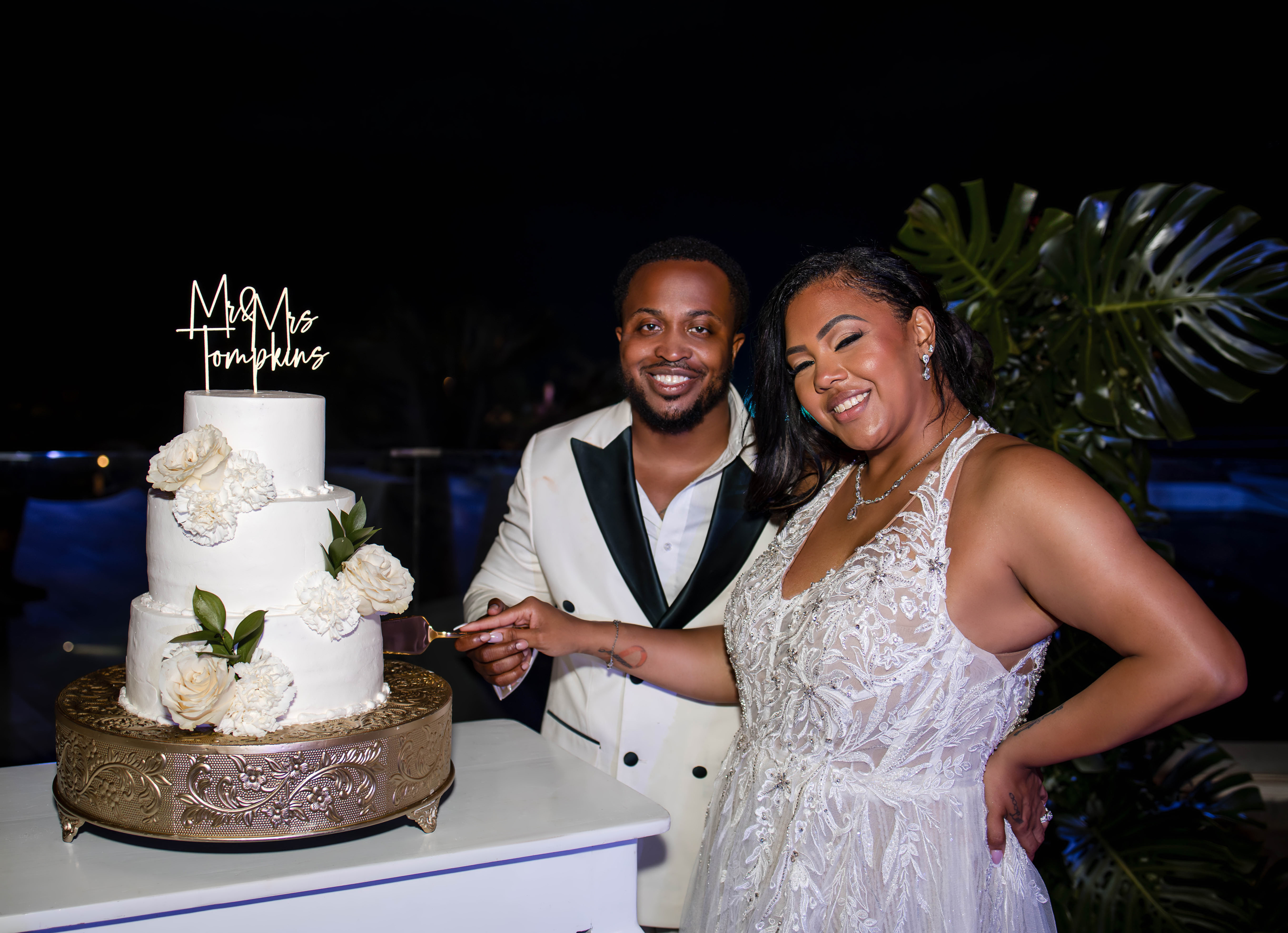Leah and Ben smile in formal wear as they cut a white wedding cake at a nighttime destination wedding.