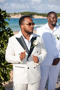 Two men in white suits by the beach, one with sunglasses and a boutonnière, at a destination wedding.