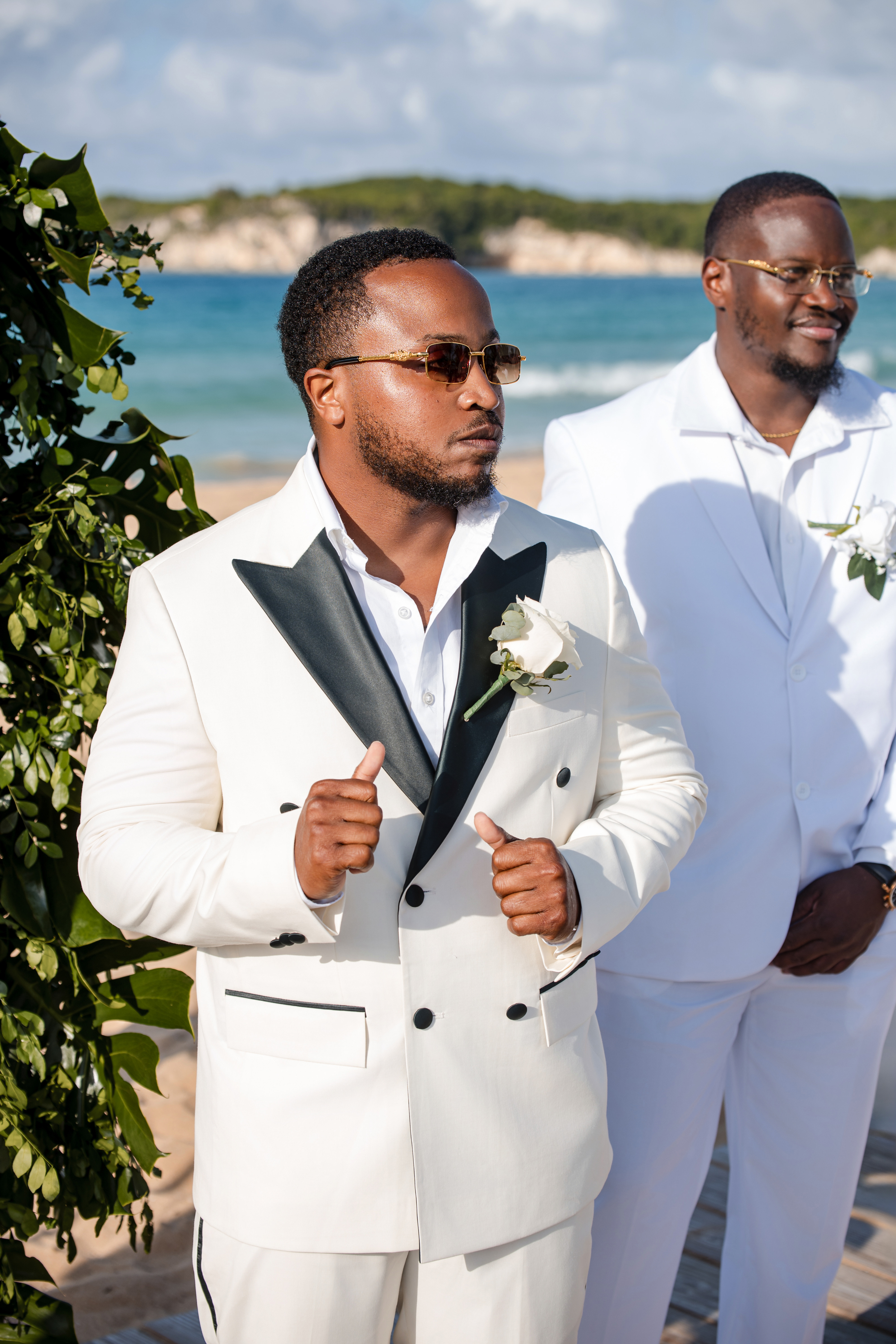 Two men in white suits by the beach, one with sunglasses and a boutonnière, at a destination wedding.