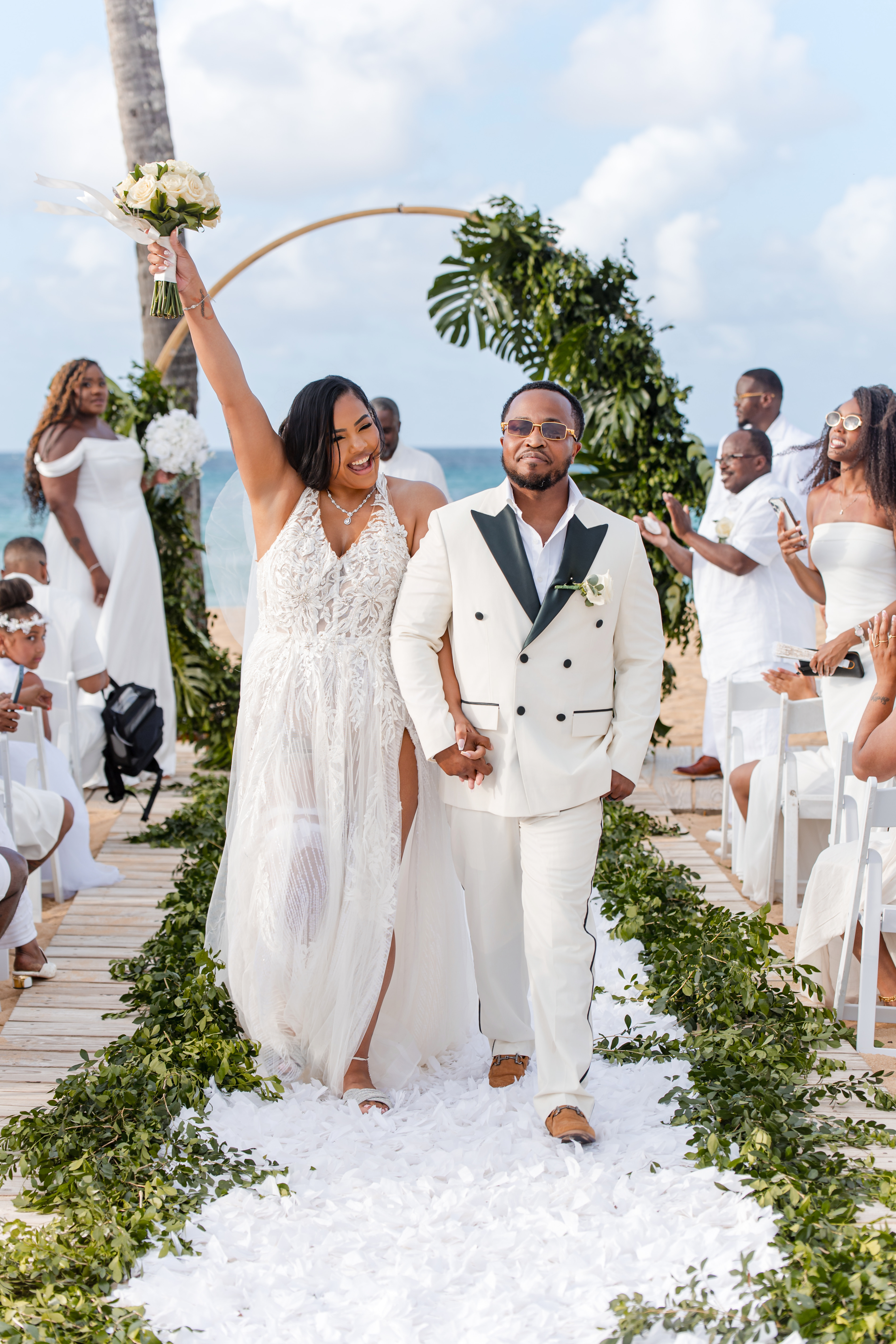 Leah and Ben walk down the aisle at their destination wedding, guests in white lining the outdoor path.