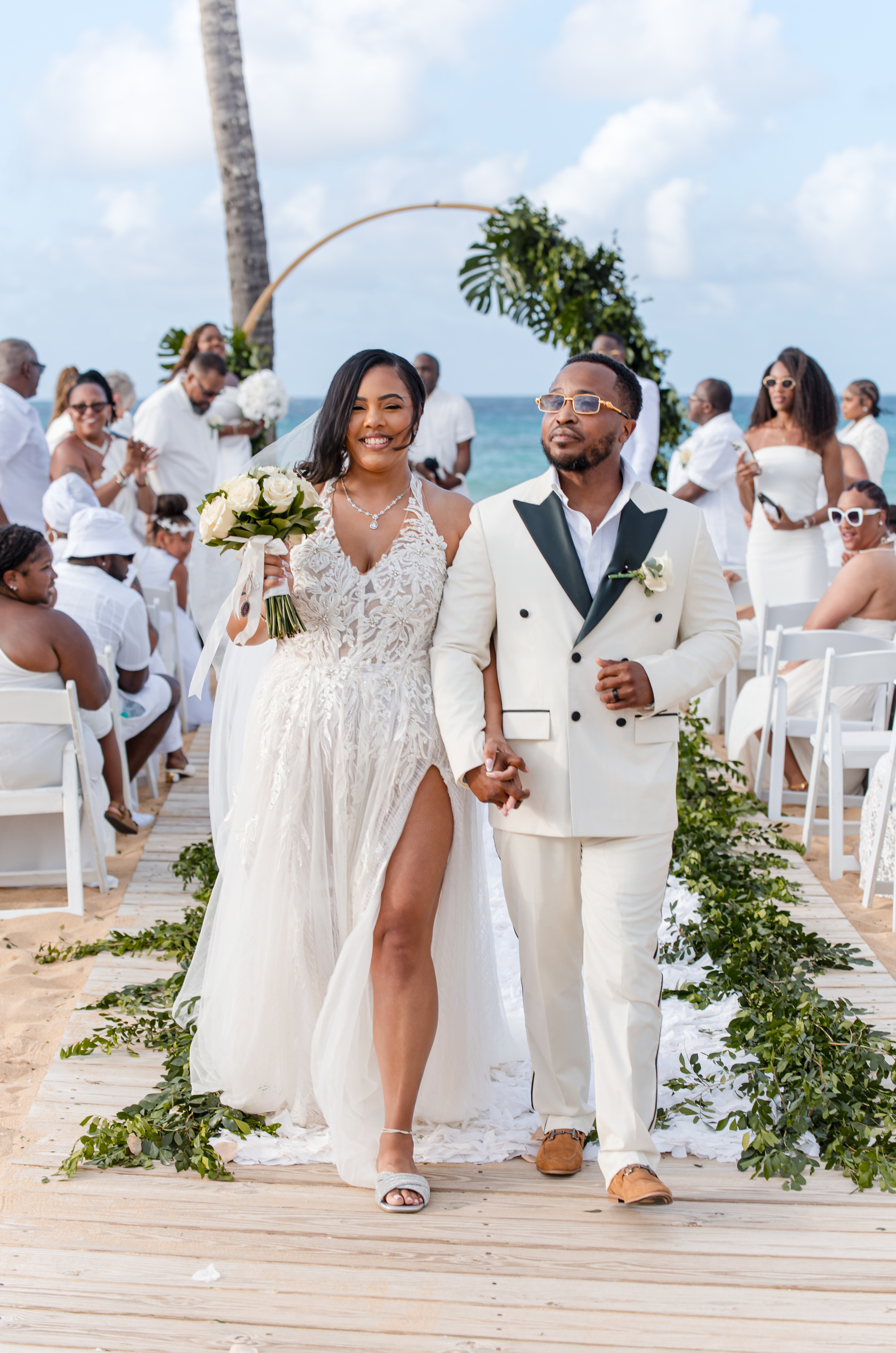 Leah and Ben walk down the aisle at their destination wedding on the beach, guests in white attire.