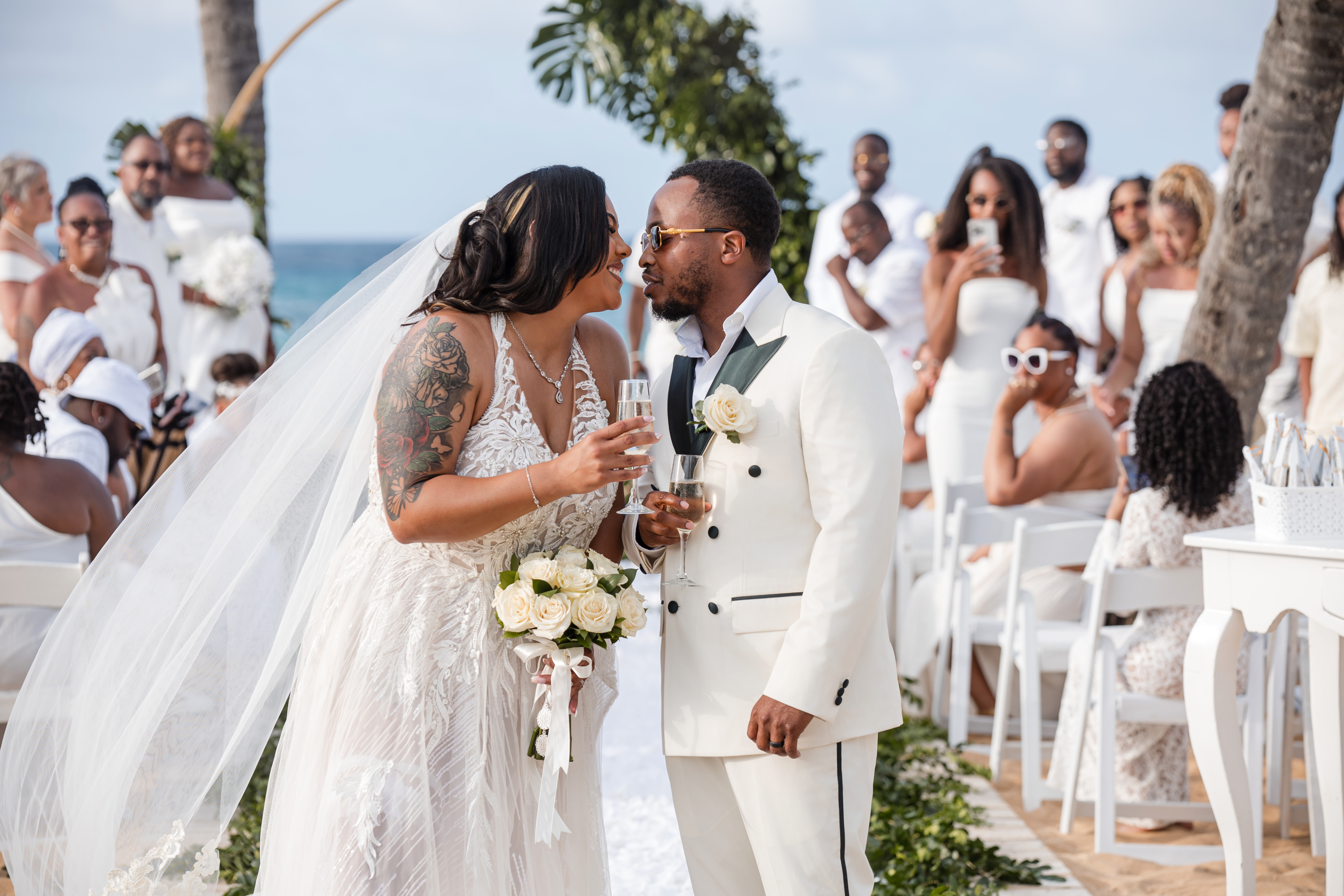 Leah and Ben in white, smiling and toasting with glasses at an outdoor destination wedding ceremony.