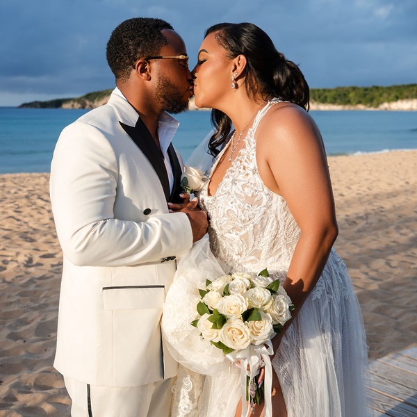 Leah and Ben share a kiss in wedding attire on a sandy beach, holding white roses at their destination wedding.