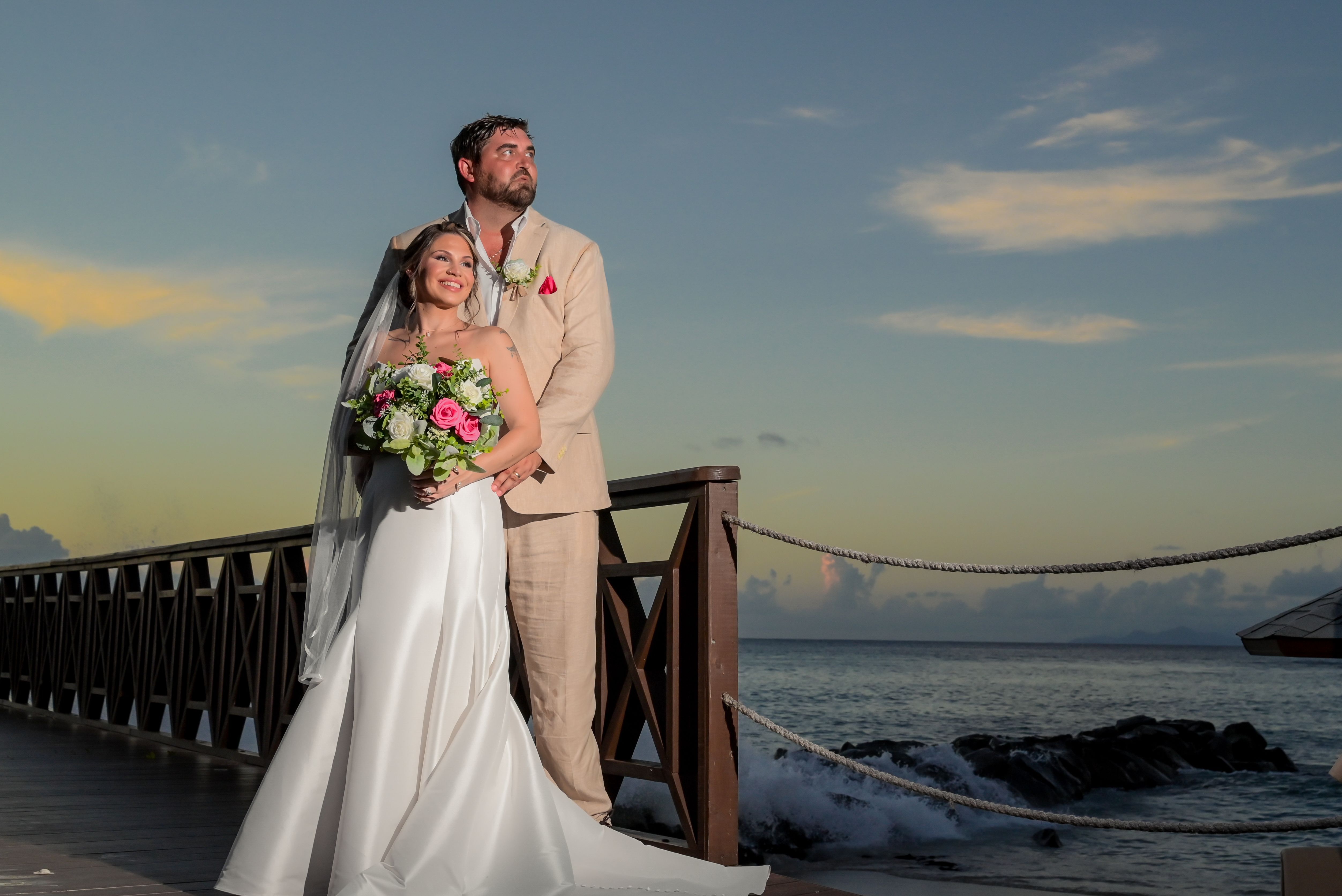 James and Brianna posing on a boardwalk by the ocean at sunset, holding a bouquet at their wedding.