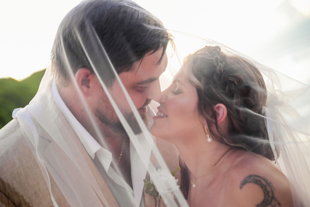 James and Brianna smile together outdoors beneath a wedding veil, enjoying their destination wedding.