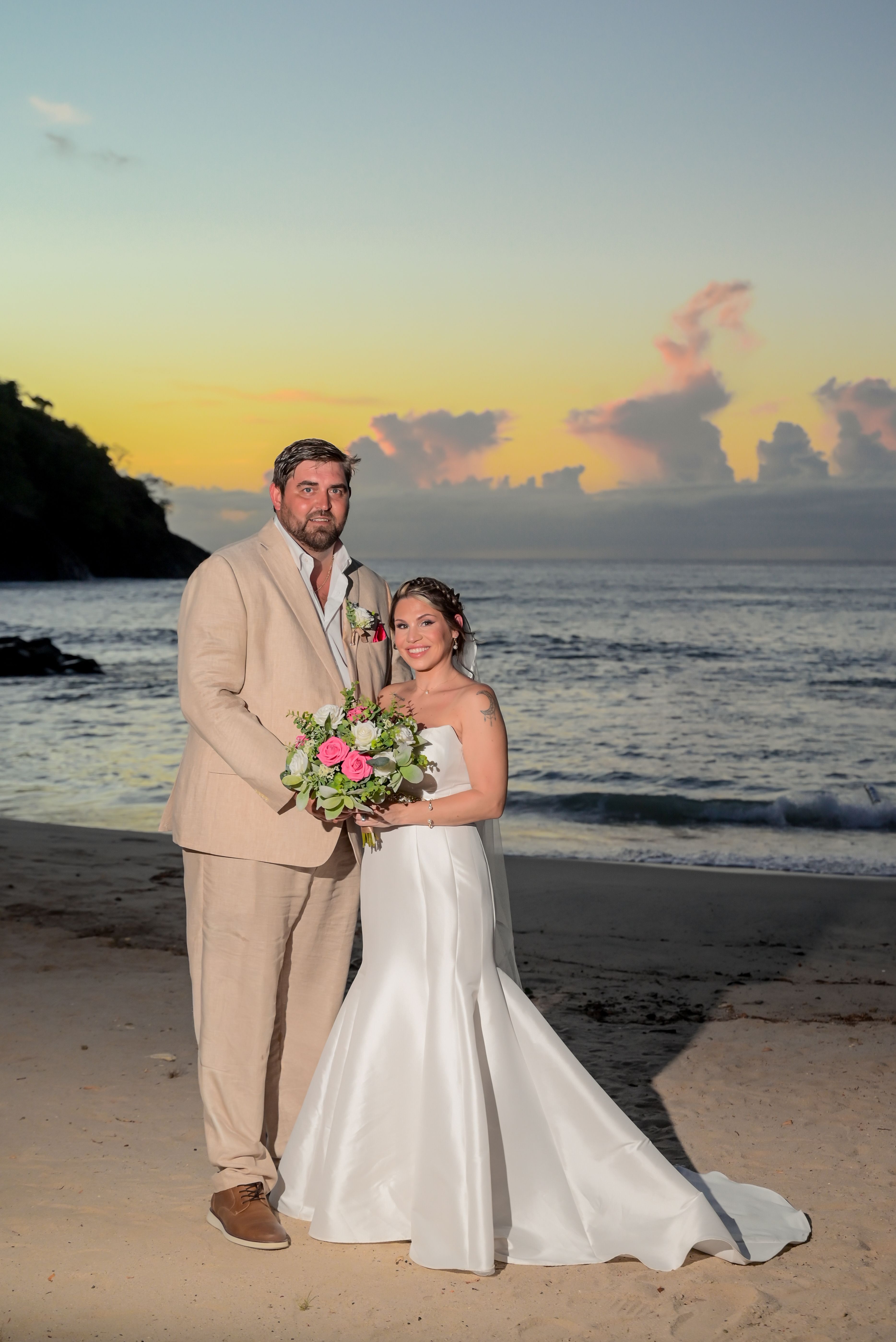 James and Brianna posing on a beach at sunset during their destination wedding, ocean in the background.