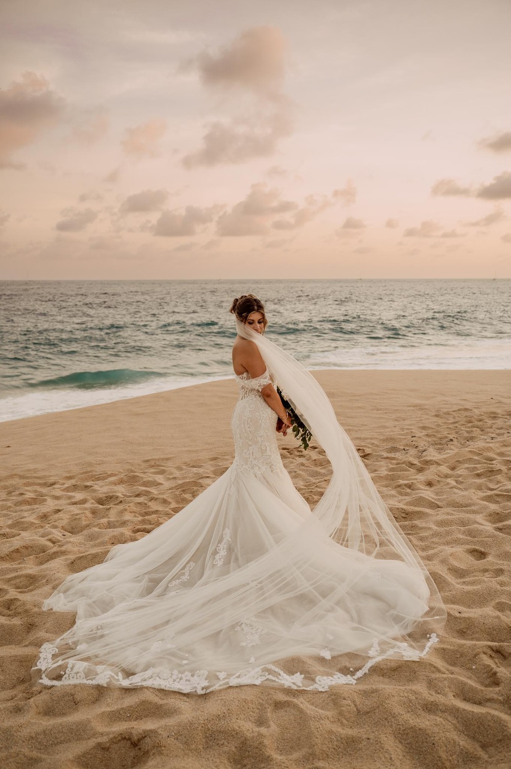 A bride in a white gown stands on a sandy beach by the ocean, perfect for a destination wedding.