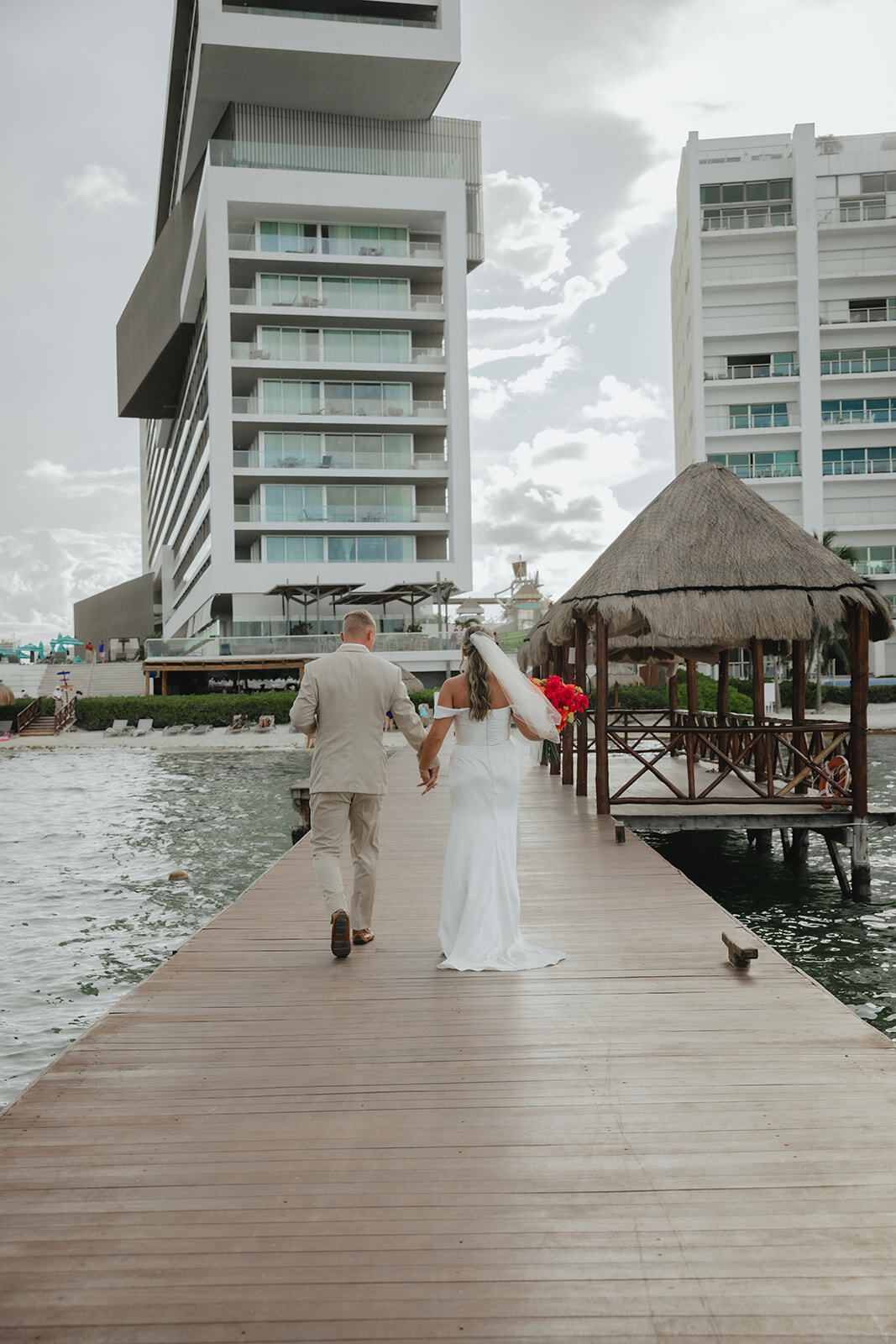 Kaitlyn and Chad stroll hand in hand on a dock by modern buildings at their destination wedding.