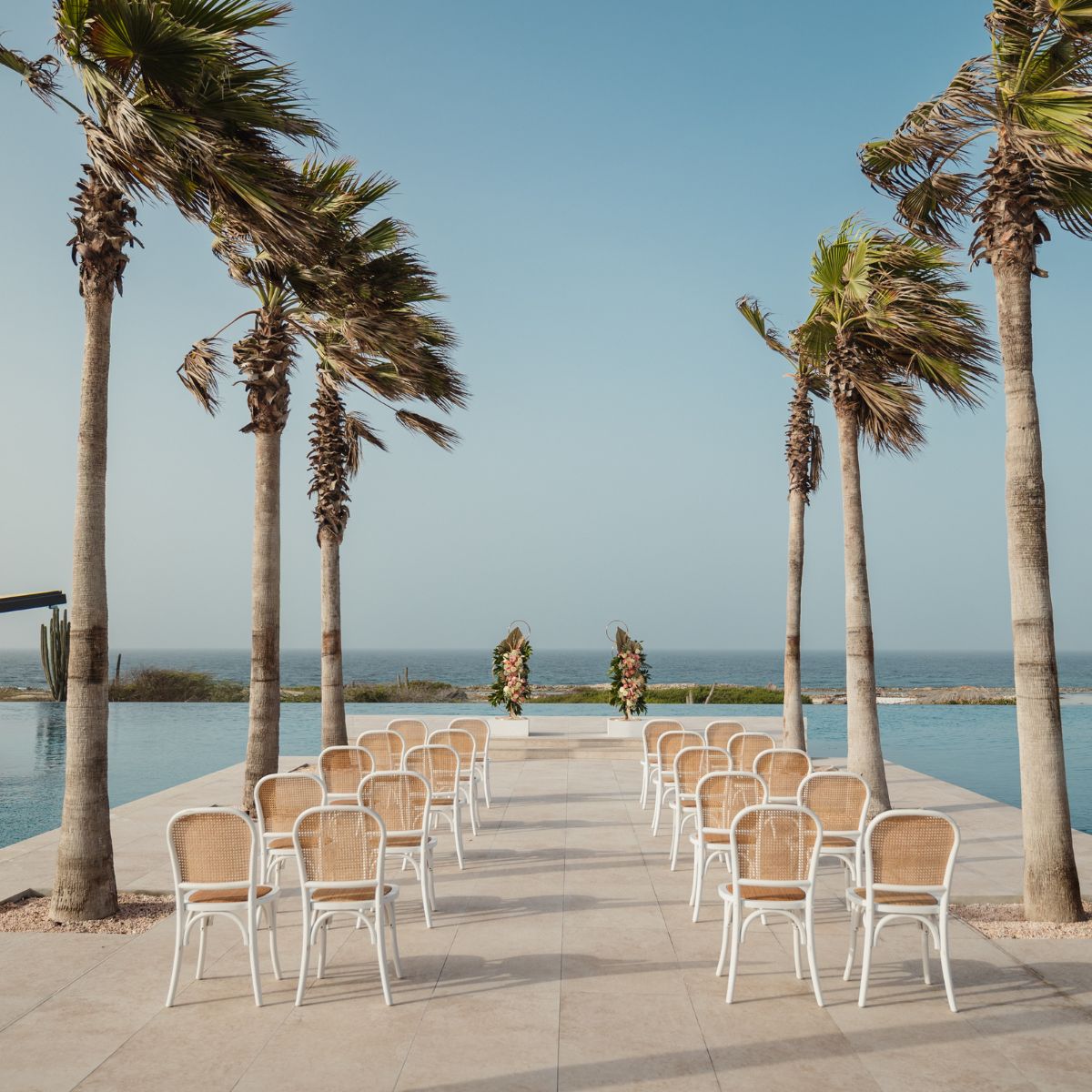 Rows of white chairs face a floral arch by a pool, palm trees and ocean create a dreamy destination wedding scene.