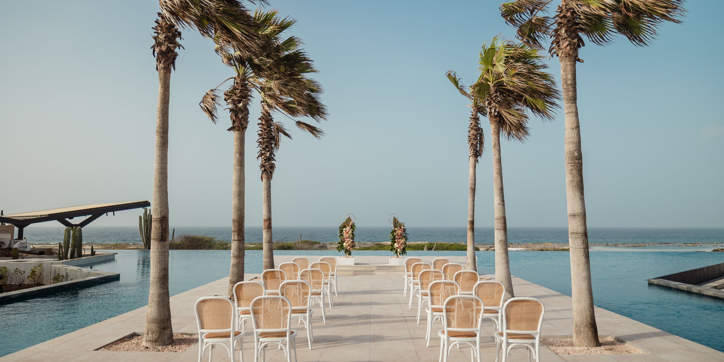 Destination wedding ceremony setup by the ocean, featuring chairs, palm trees, and floral decorations.