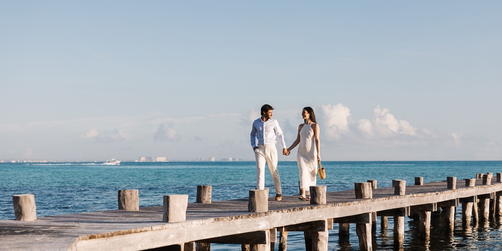 A couple walks hand in hand on a wooden pier over clear blue water at their destination wedding.