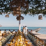 A couple stands by a set table at their destination wedding, overlooking a pier and ocean under wicker lights.