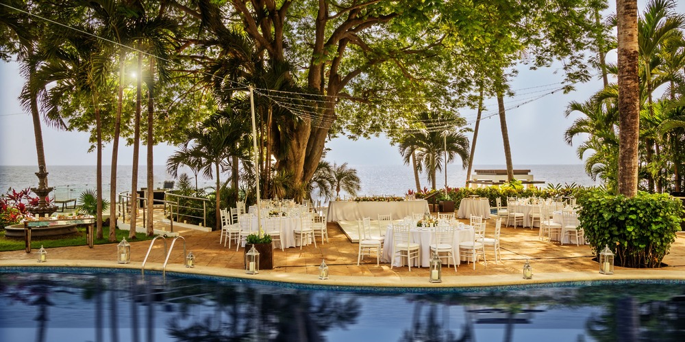Destination wedding setup with round tables by a pool, trees, and ocean view in the background.