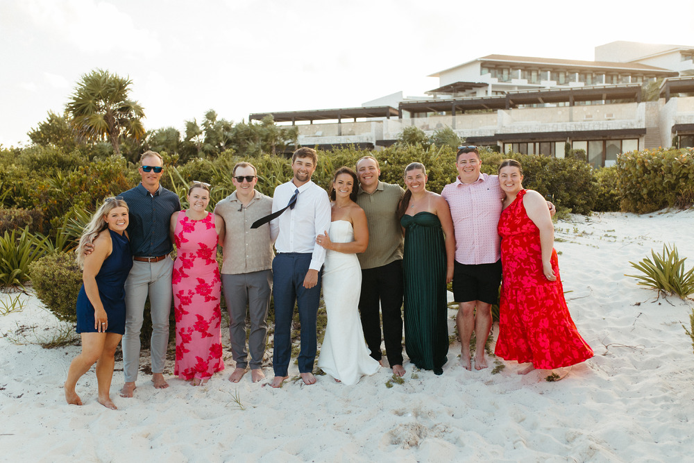 A group poses barefoot on the sandy beach at a destination wedding in front of Shirley and William.