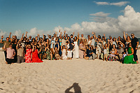 Large group at a destination wedding posing on a sandy beach beneath a clear blue sky.