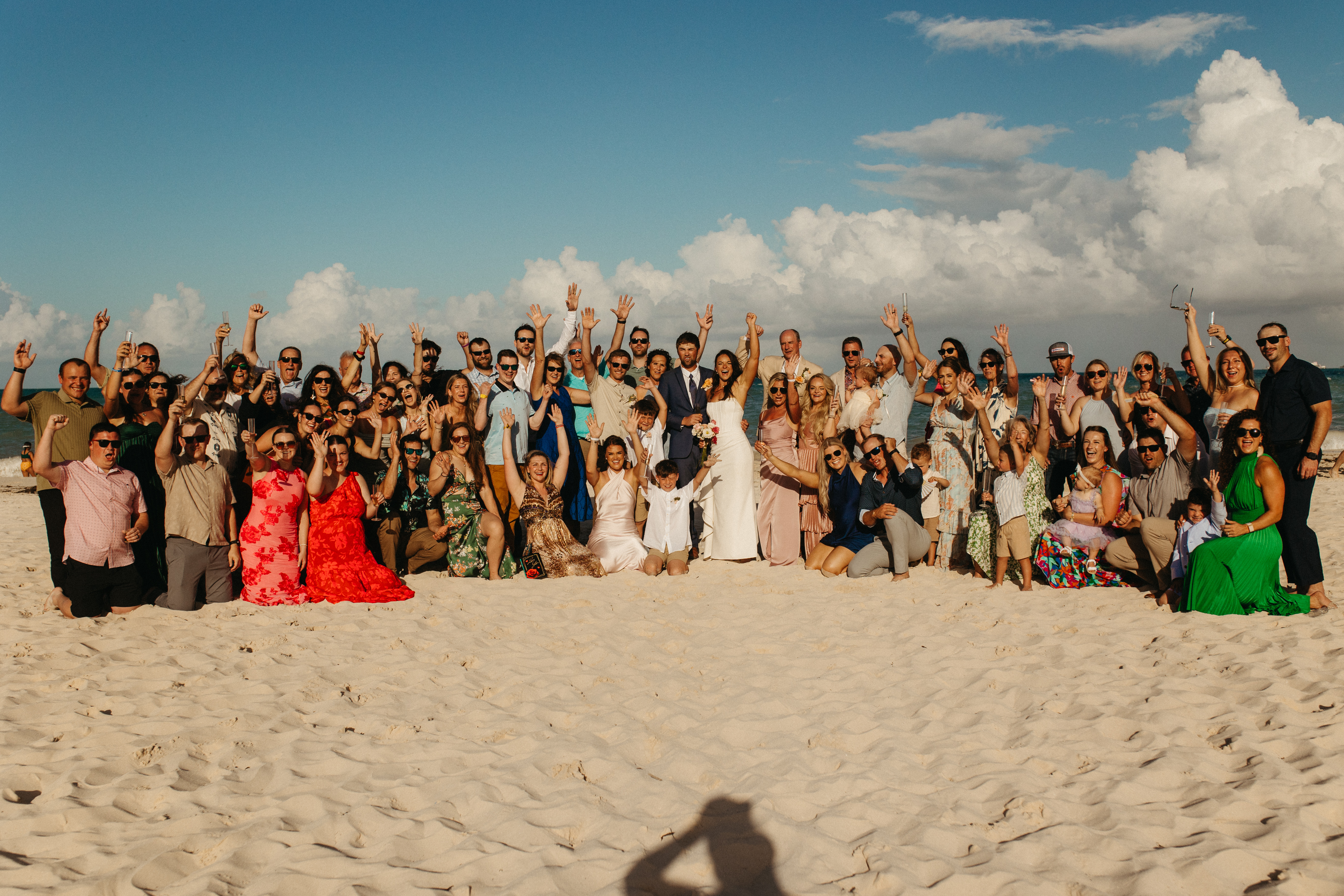 Large group at a destination wedding posing on a sandy beach beneath a clear blue sky.