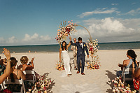 Shirley and William walk down a beach wedding aisle beneath a floral arch on a bright, sunny day.