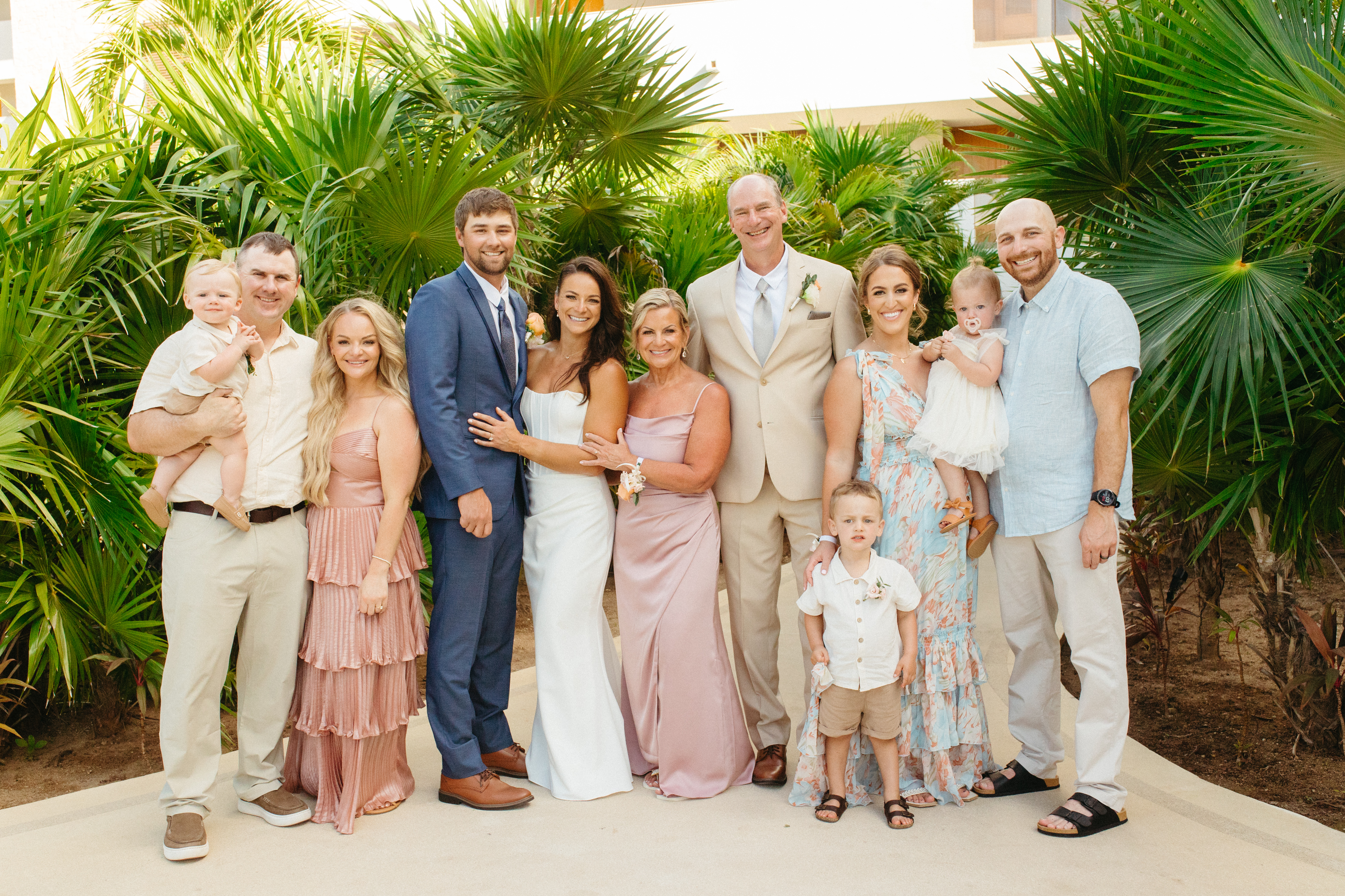 Eleven people pose outdoors by lush tropical plants, dressed for a destination wedding celebration.