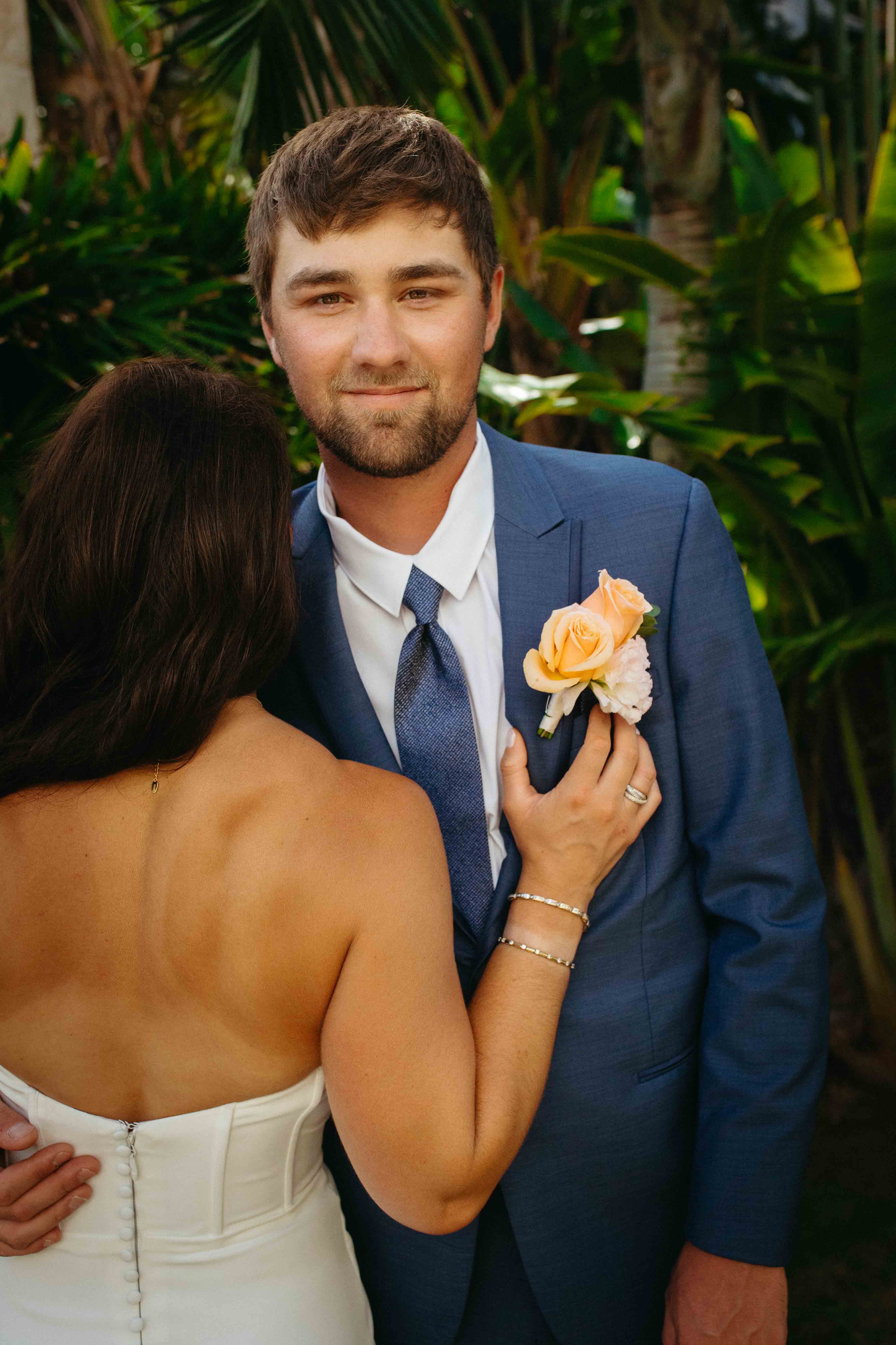 At a destination wedding, a woman in white embraces a man in a blue suit with peach rose boutonniere.