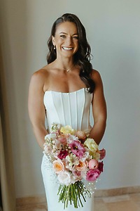 Bride in a "Shirley and William (Copy)" white dress smiles at a destination wedding with vibrant bouquet.