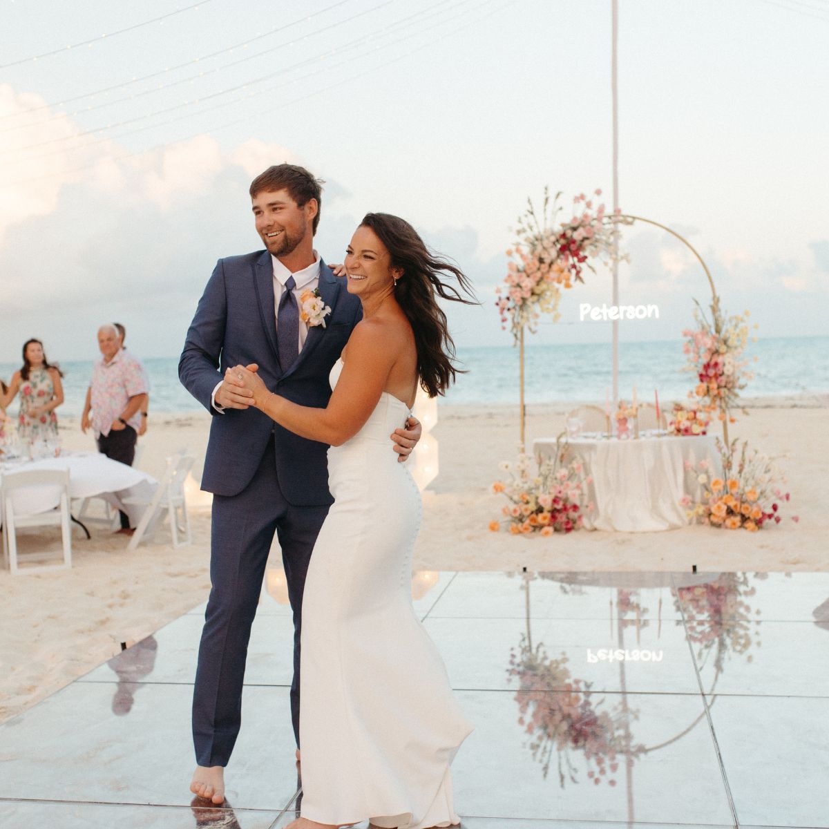 Bailey and Justin share a dance at their destination wedding, with an arch and table on the beach behind them.