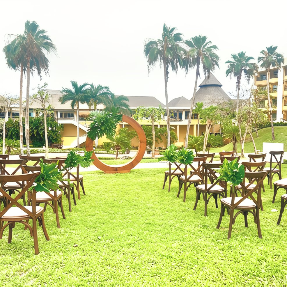 Destination wedding ceremony with wooden chairs, lush greenery, and a round floral arch on the lawn.