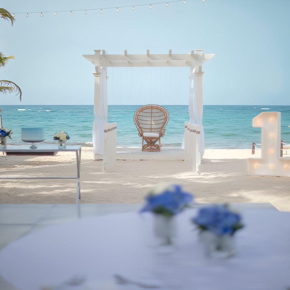White pergola with wicker chair on sandy beach, ocean behind, decorated for a destination wedding.