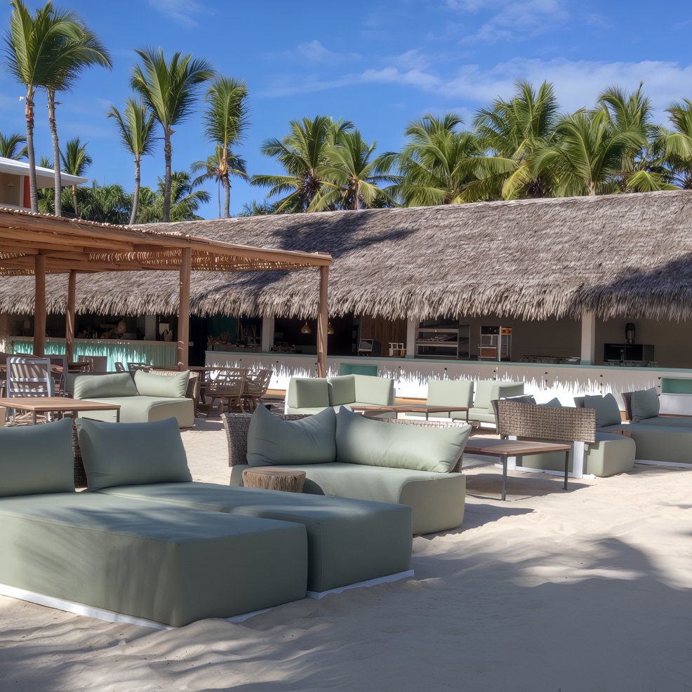 Outdoor lounge with cushioned seats on sandy beach, ideal for a destination wedding by palm trees and bar.