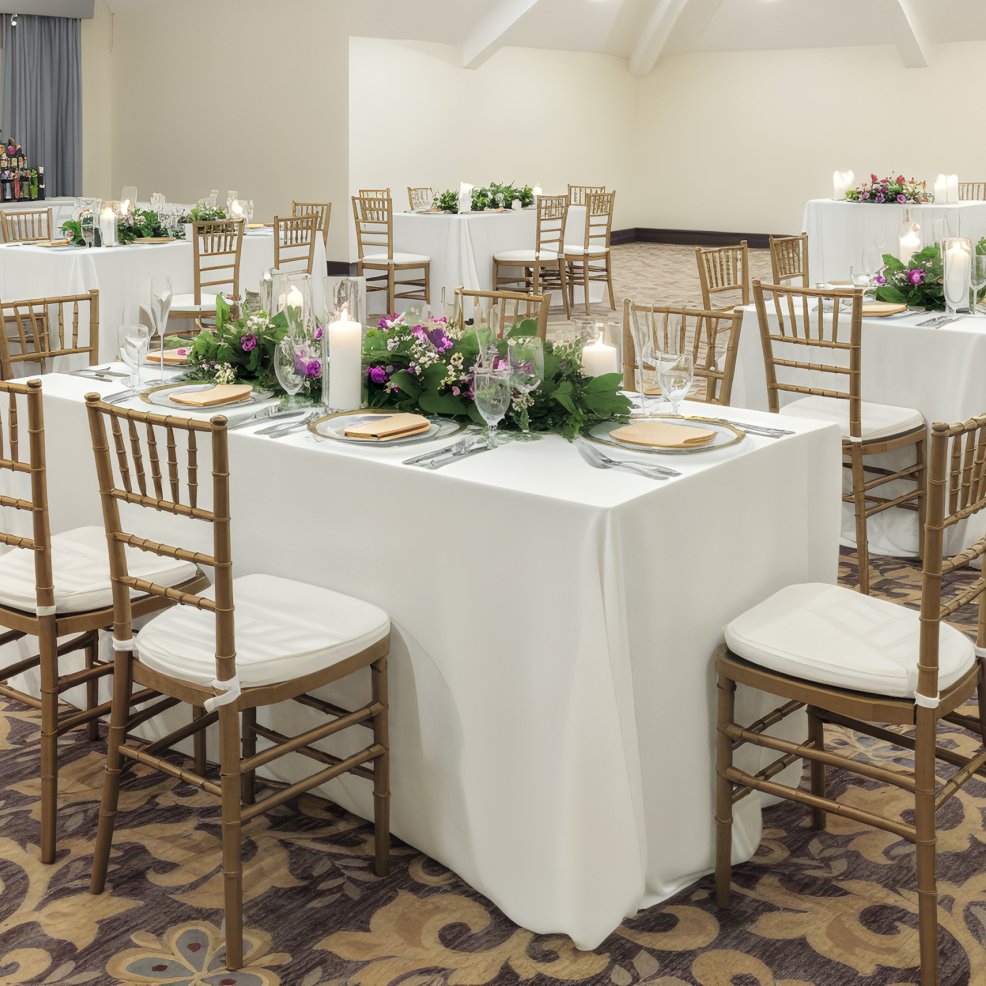 Elegant wedding dining tables with white linens, floral centerpieces, and wooden chairs at a destination.
