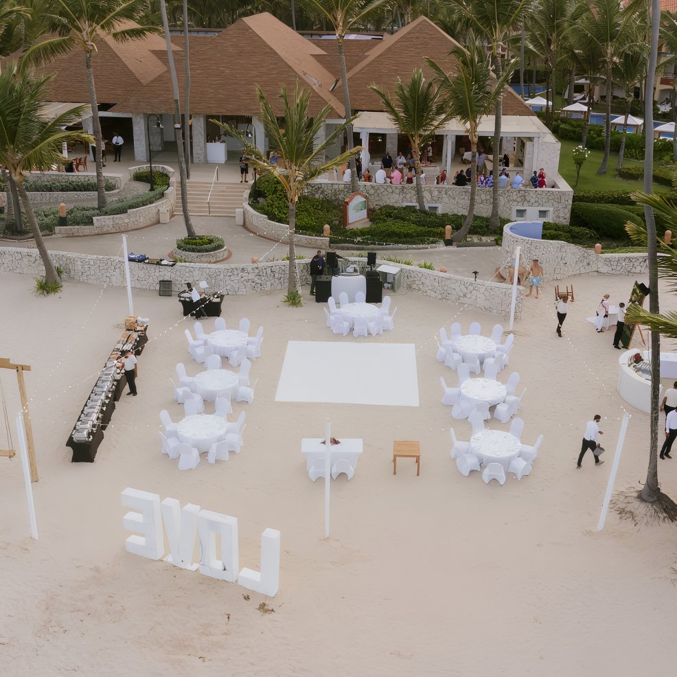 Destination wedding setup on the beach with round tables, white chairs, and large "LOVE" letters displayed.