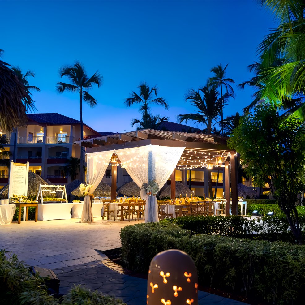 Destination wedding setup outdoors at dusk with string lights, white drapes, and lush palm trees.