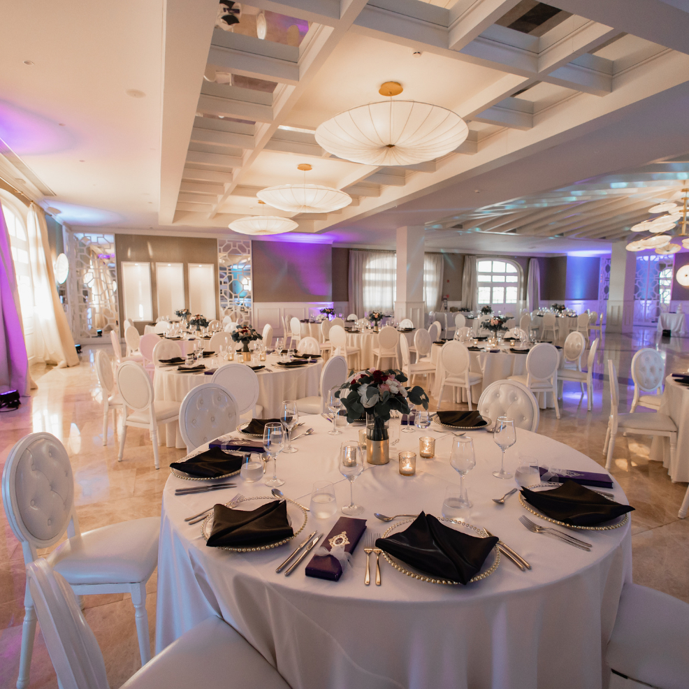 Elegant banquet hall set for a destination wedding, with round tables, white chairs, and purple lighting.