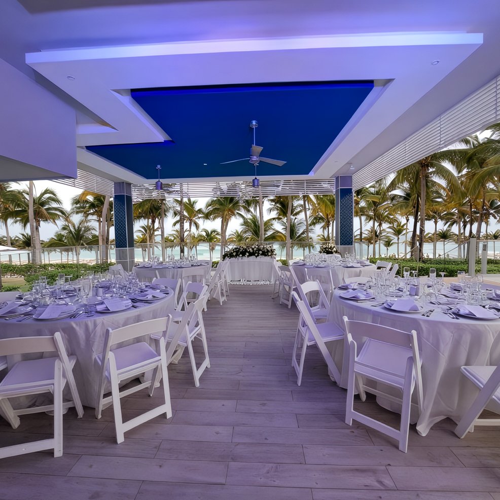 Destination wedding reception outdoors with round tables, white chairs, and palm trees in the background.