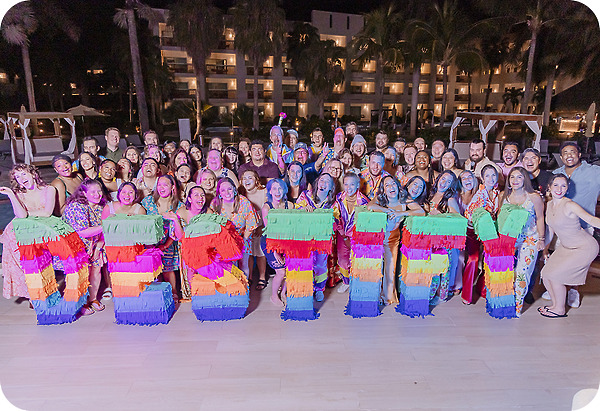A large group at a destination wedding poses at night outdoors, holding colorful piñatas spelling “FIAMMA.”.