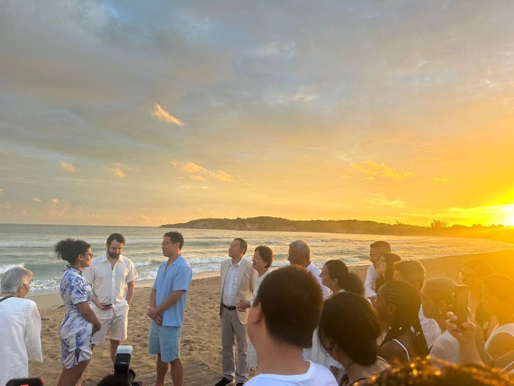 Brianna and Aszah stand face to face at their destination wedding on a sunset beach, friends gathered.