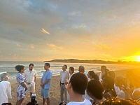Brianna and Aszah stand face to face at their destination wedding on a sunset beach, friends gathered.