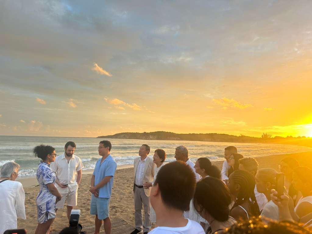 Brianna and Aszah stand face to face at their destination wedding on a sunset beach, friends gathered.