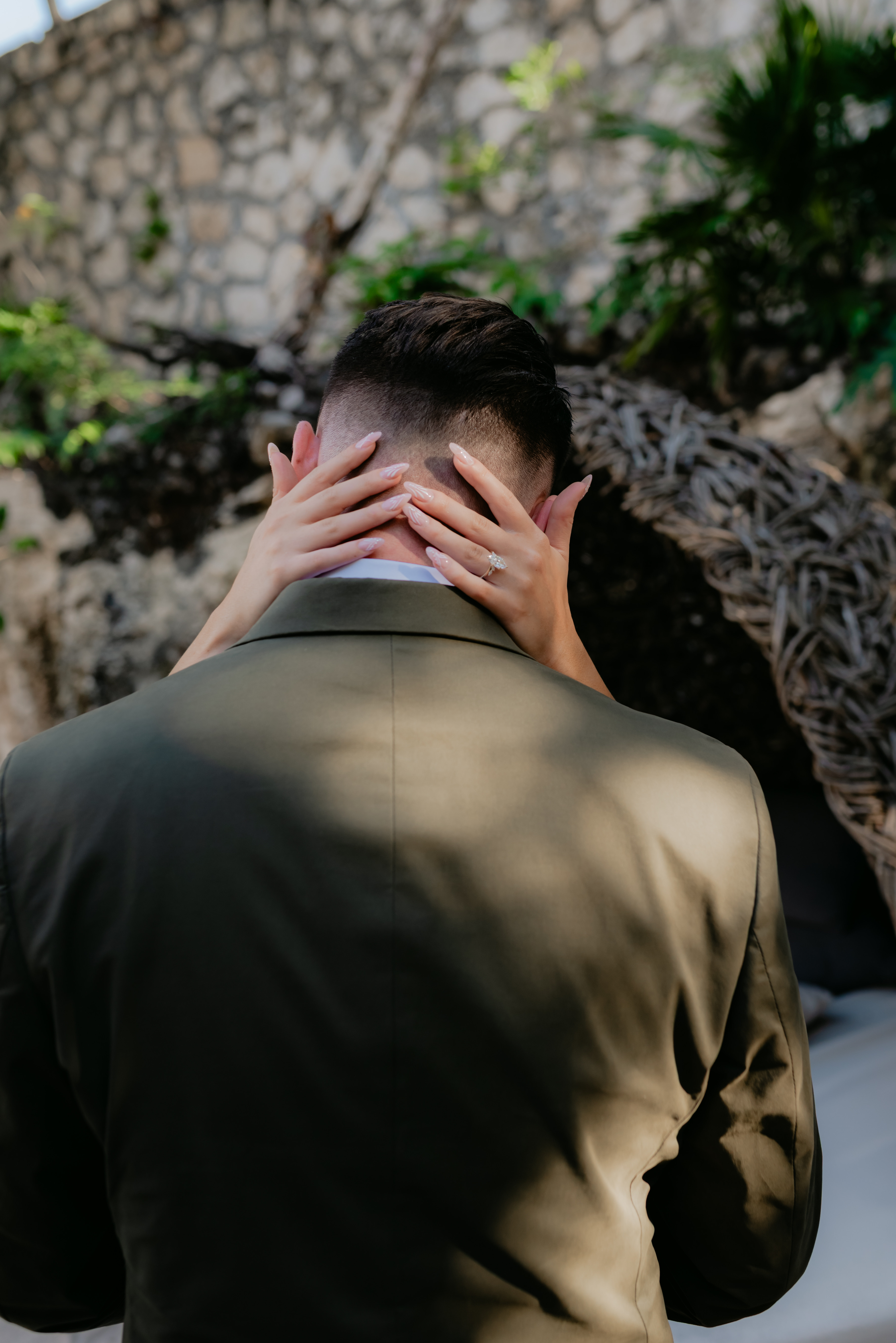 Person in a suit embraced from behind at a wedding, ring visible on hand.