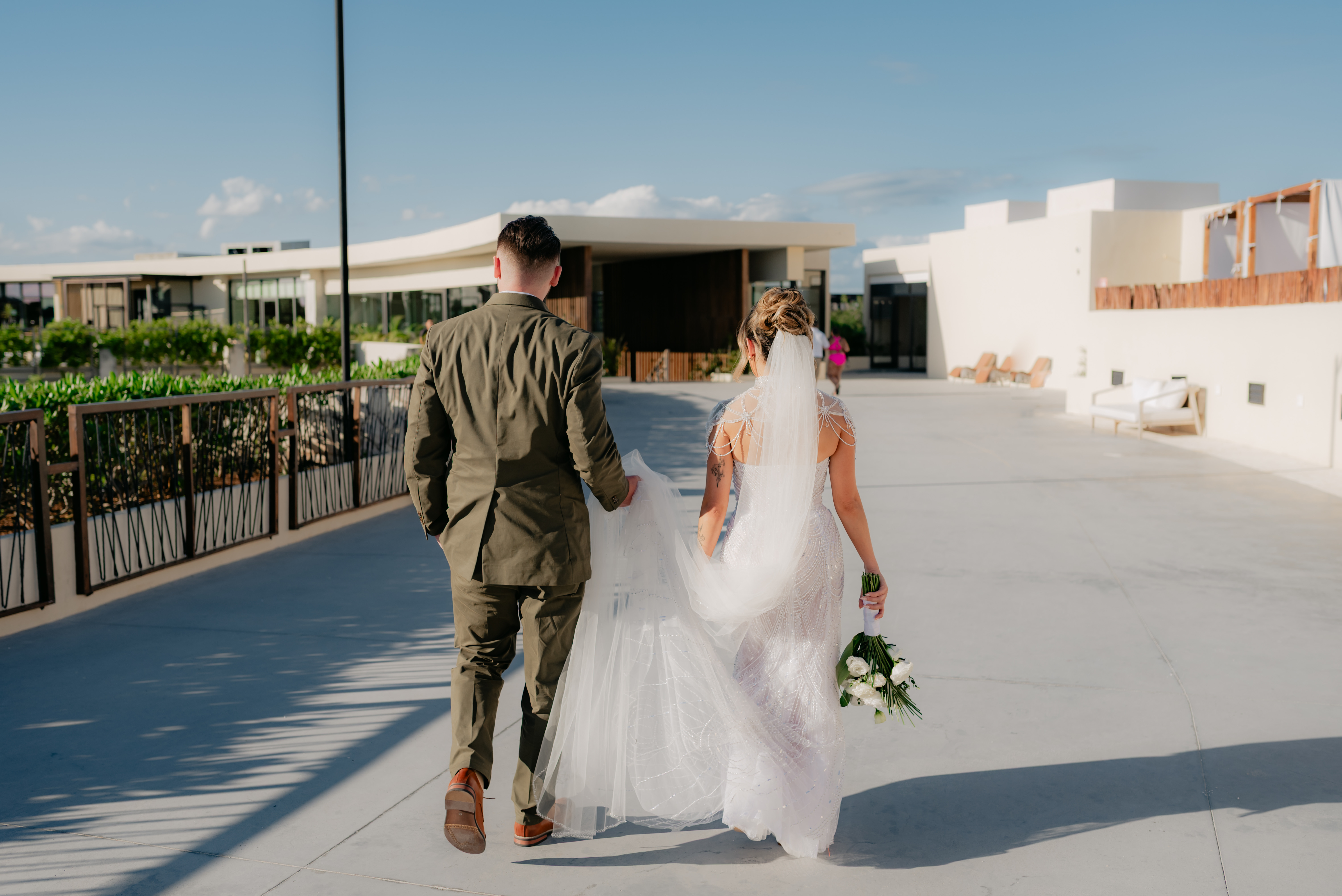Bride and groom walk outdoors on a sunny day during their destination wedding; bride holds bouquet.