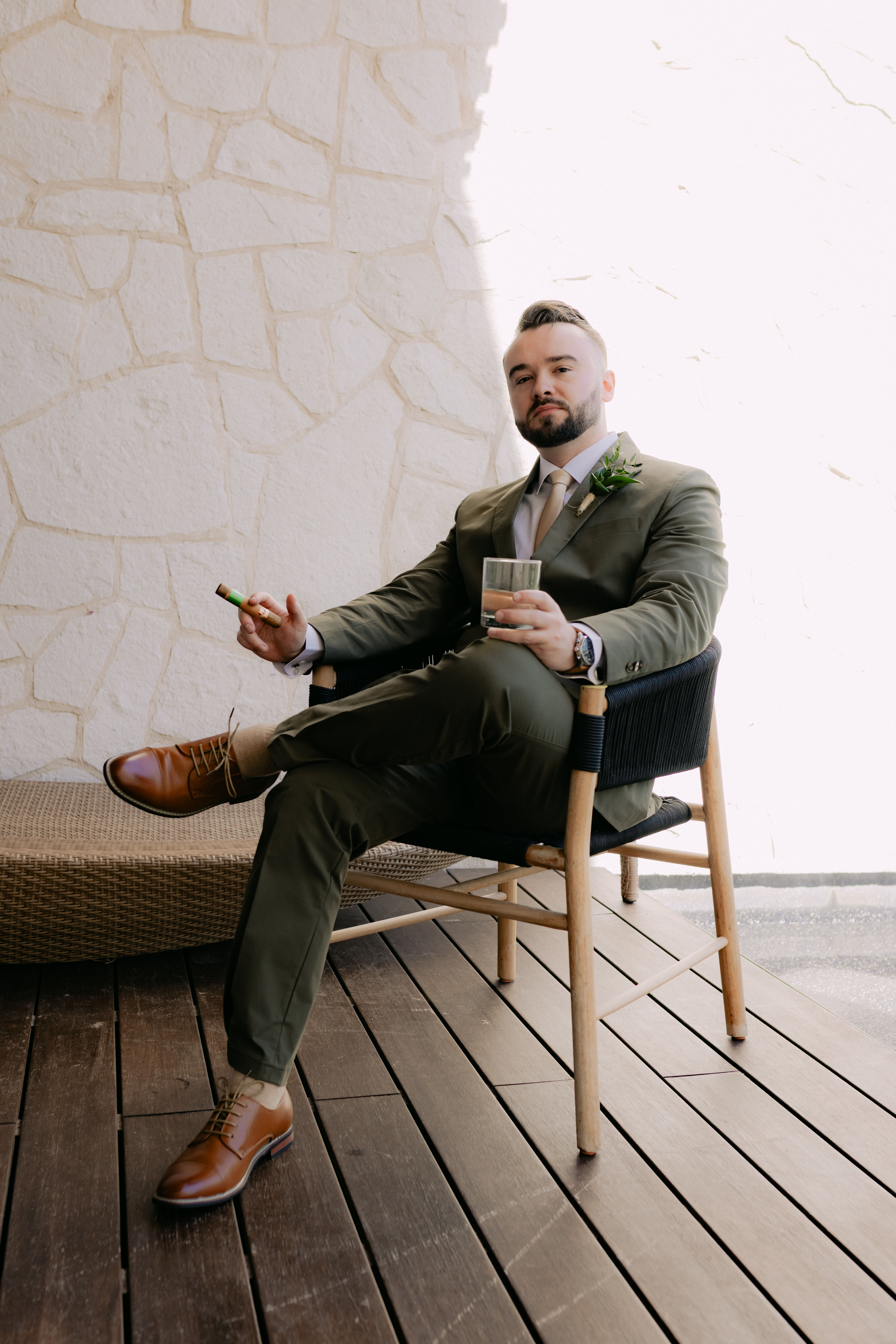 Man in suit on chair with drink and cigar by stone wall at destination wedding; Shirley and William.