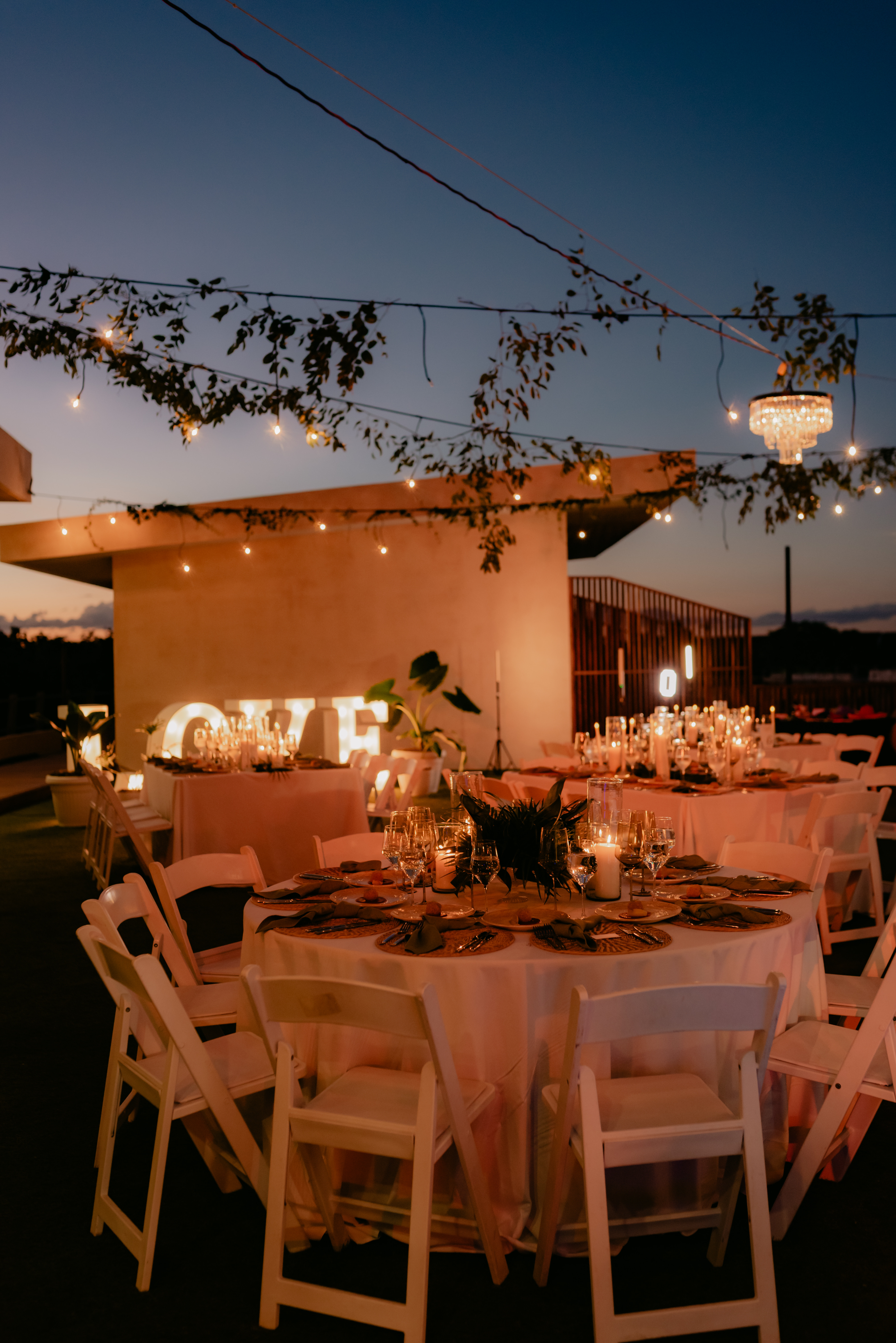 Round tables with white chairs set for dinner under string lights at a destination wedding.
