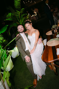 At a lush venue, the bride in white sits on the groom’s lap during their destination wedding.