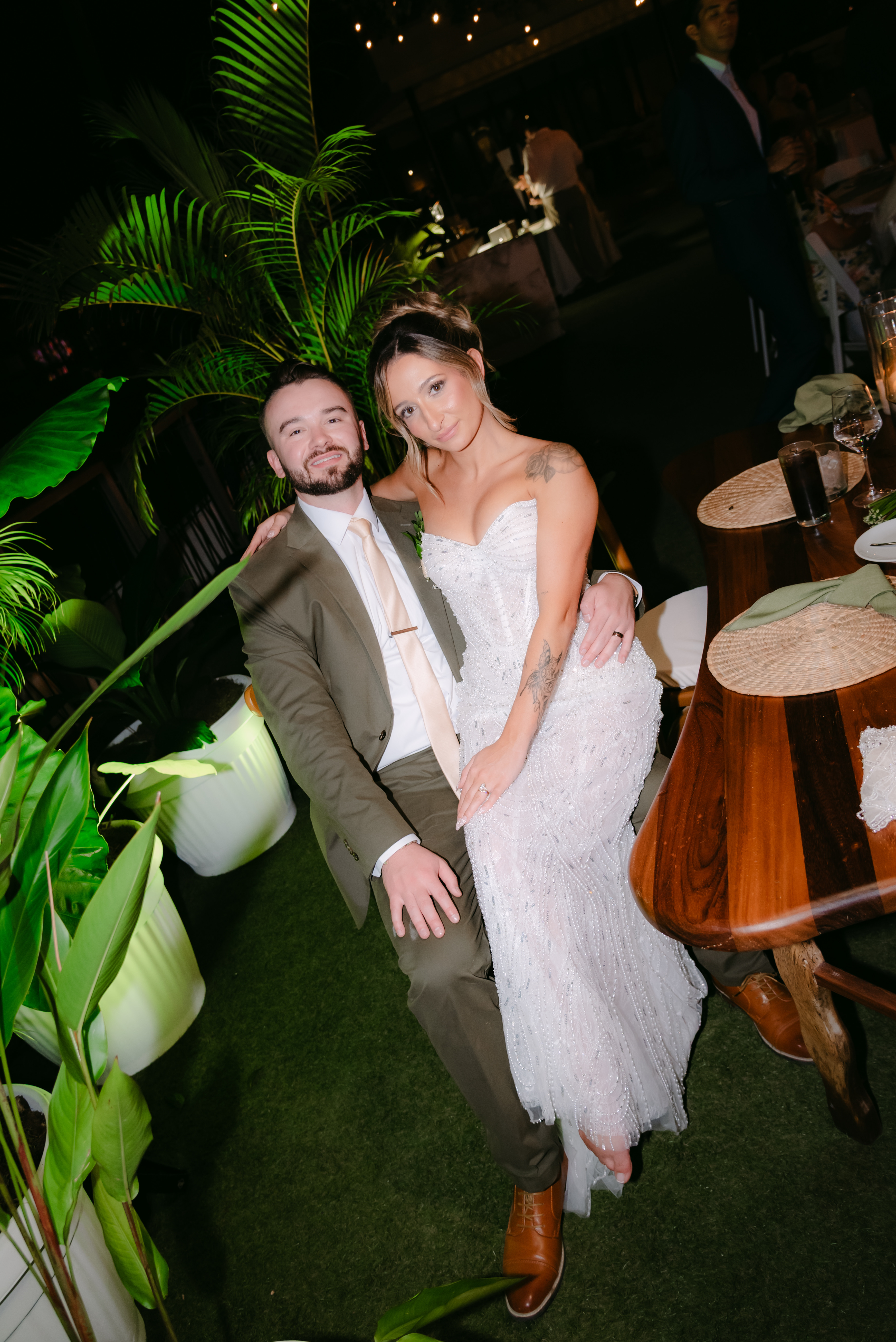 At a lush venue, the bride in white sits on the groom’s lap during their destination wedding.