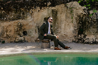 Man in a suit by a rock wall and pool, holding a drink at a destination wedding. Product: Shirley and William.