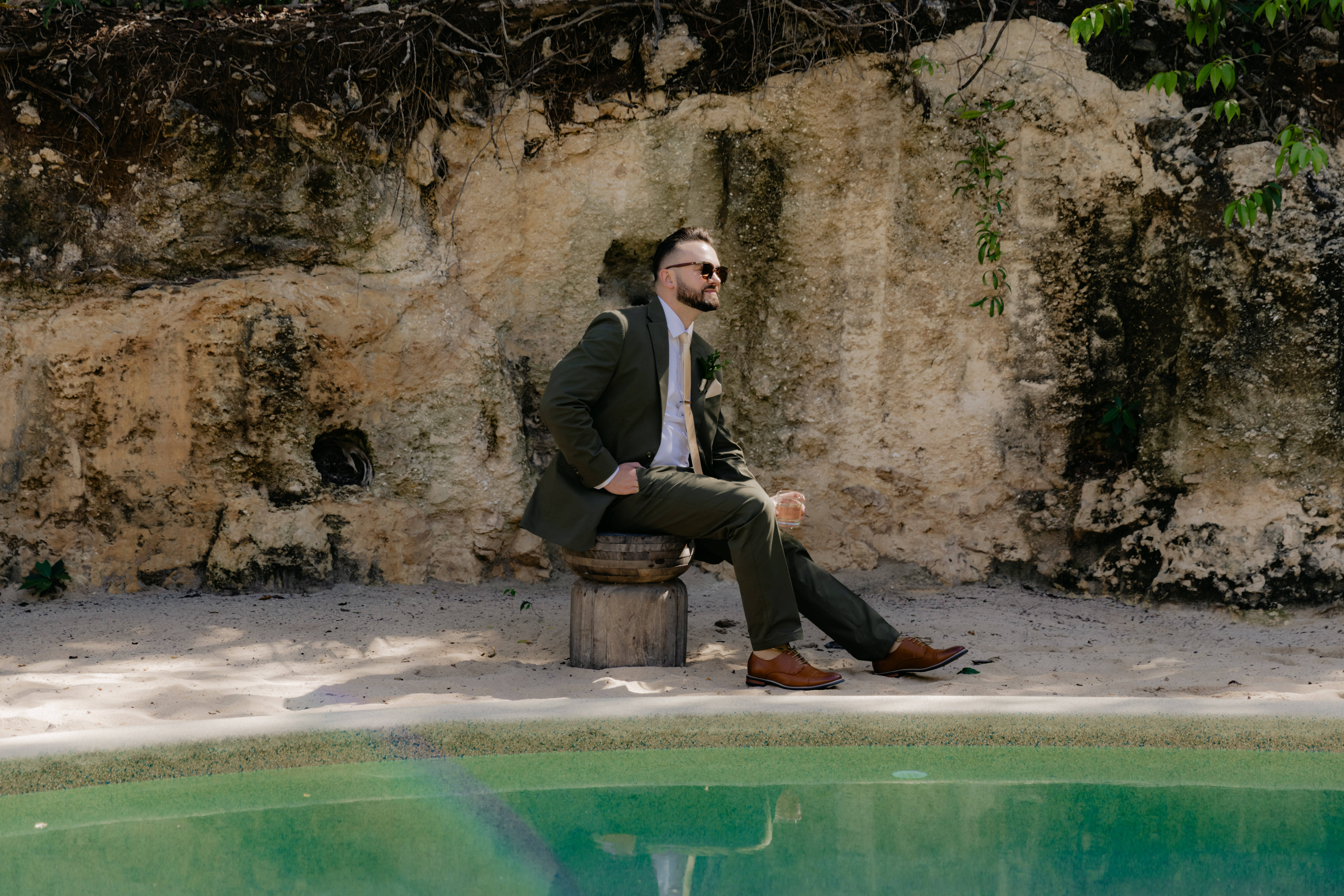 Man in a suit by a rock wall and pool, holding a drink at a destination wedding. Product: Shirley and William.