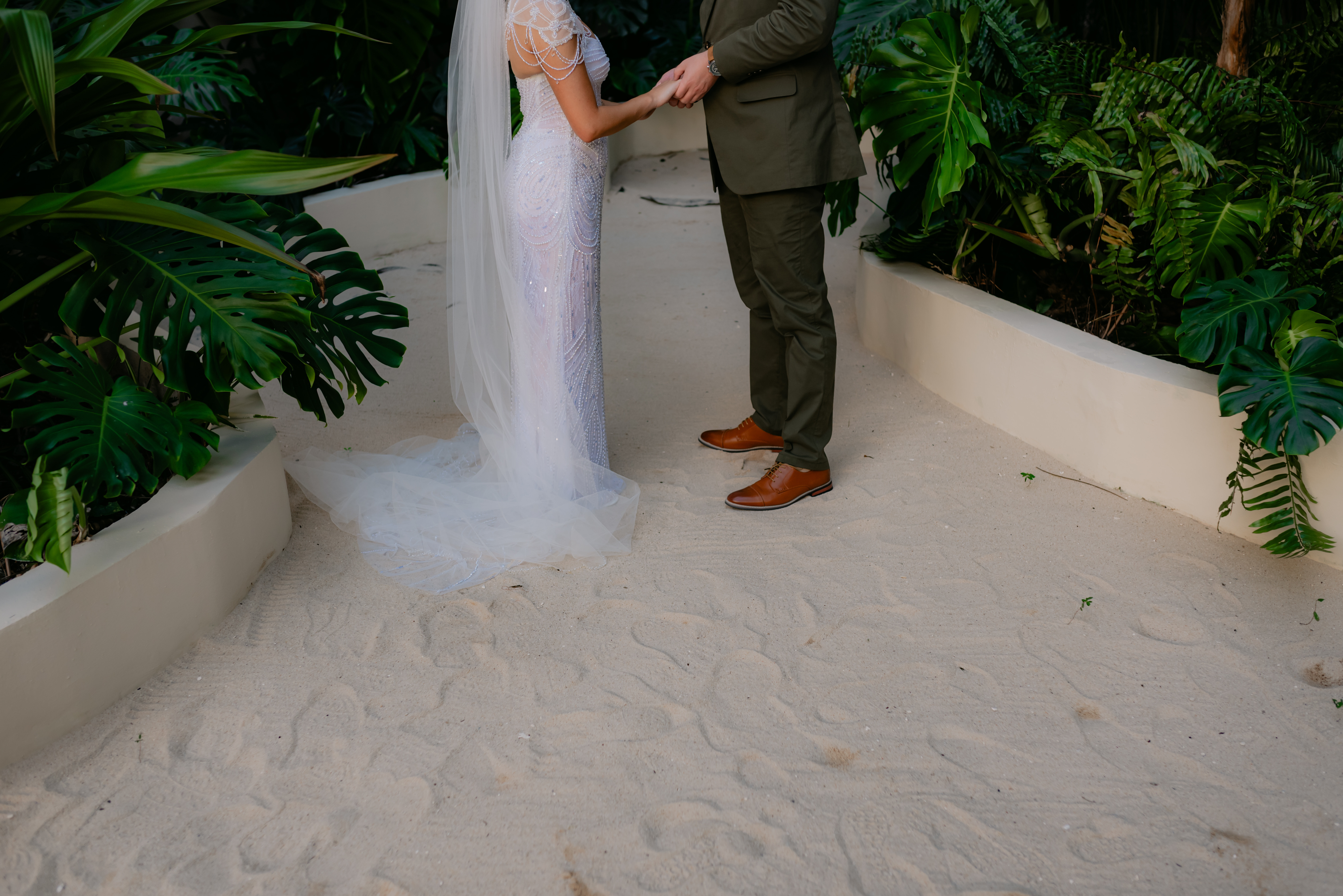 Shirley and William holding hands on a sandy path at their destination wedding, lush greenery all around.