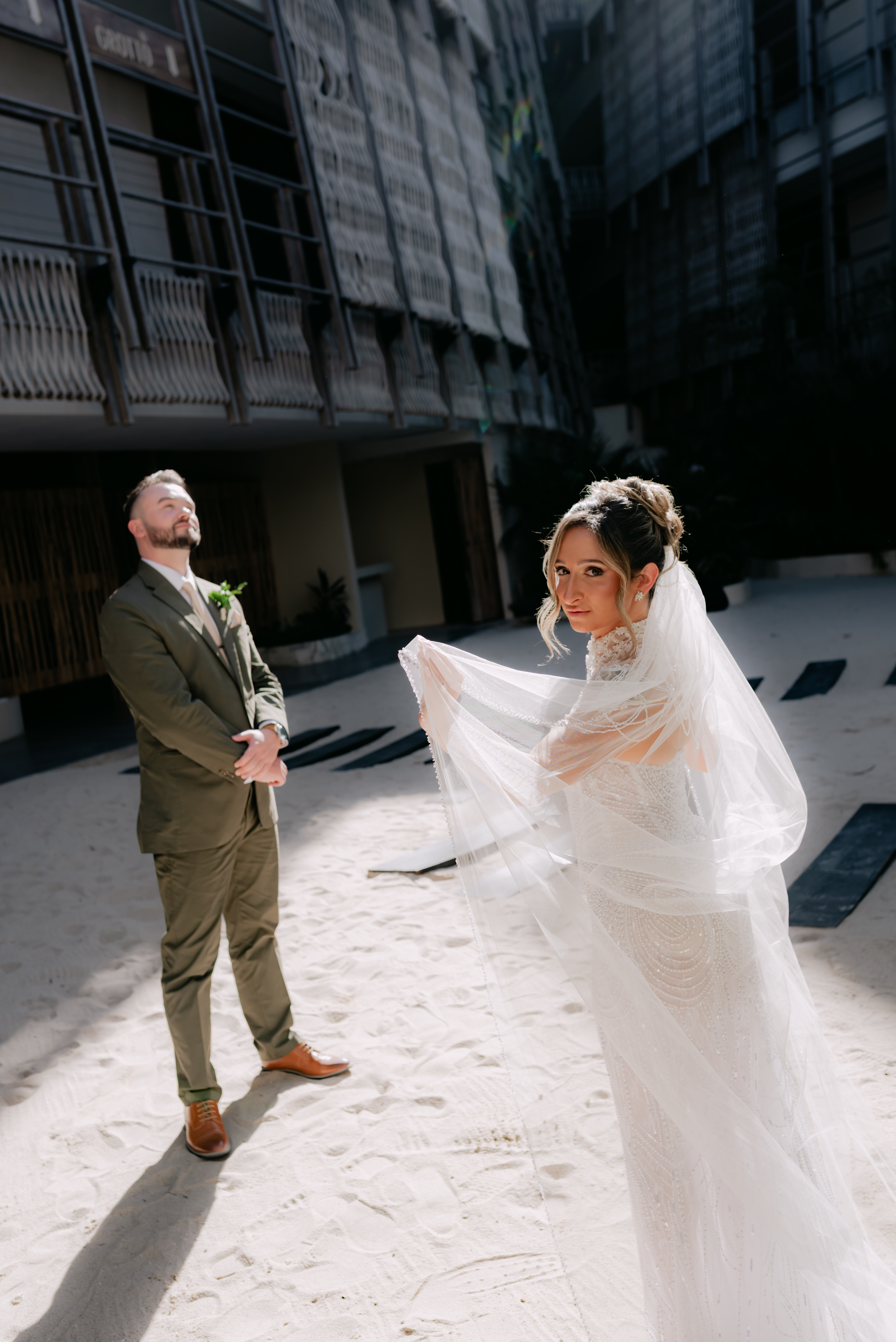 Bride in white and groom in green suit stand together in a sunlit courtyard at their destination wedding.