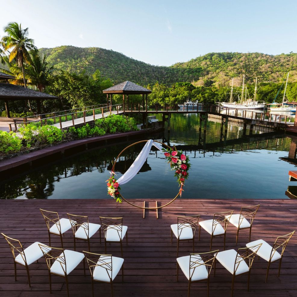 Destination wedding ceremony by the water, floral arch and chairs set up, mountains visible in the background.