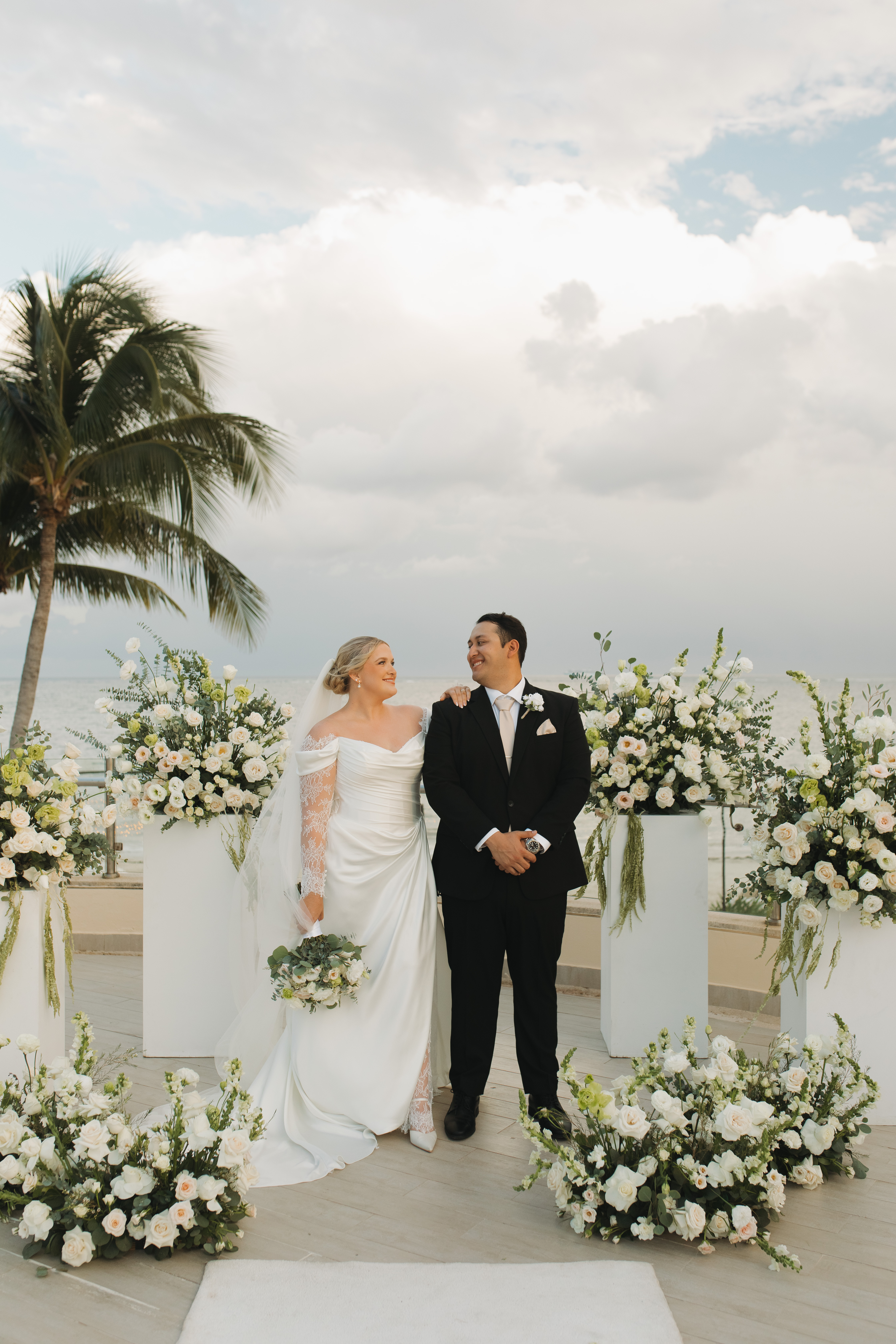 Madison and Robert pose outdoors at their destination wedding, surrounded by white flowers and palm trees.