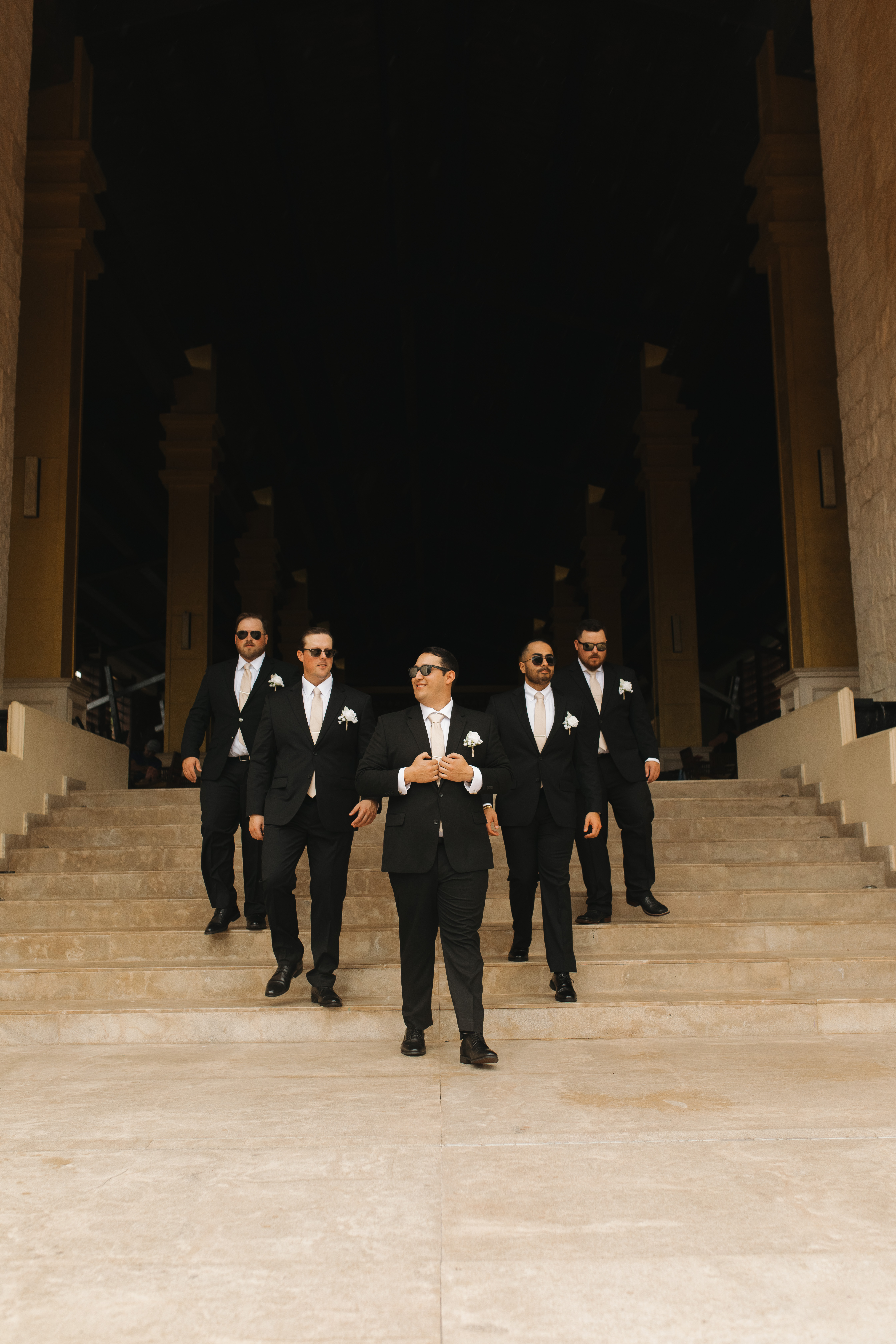 Madison and Robert descend stone steps in black suits and sunglasses at their destination wedding.